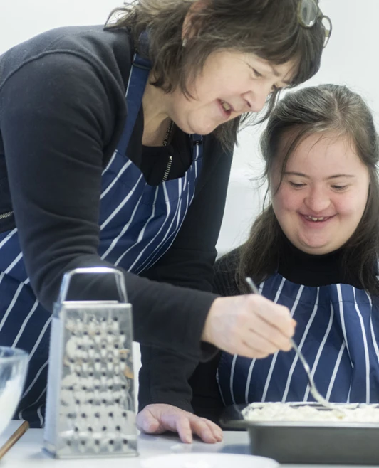 Woman and young woman with Down syndrome smiling while preparing food together in a kitchen setting, wearing striped aprons Woman and young woman with Down syndrome smiling while preparing food together in a kitchen setting, wearing striped aprons